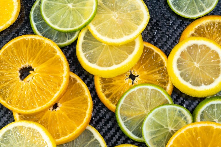 A vibrant, top-down close-up shot of multiple freshly cut citrus fruit slices arranged on a glossy, black carbon-fiber textured surface. The arrangement includes bright orange, yellow lemon, and green lime slices, showcasing glistening juice droplets, translucent pulp, and visible segment lines where they overlap.