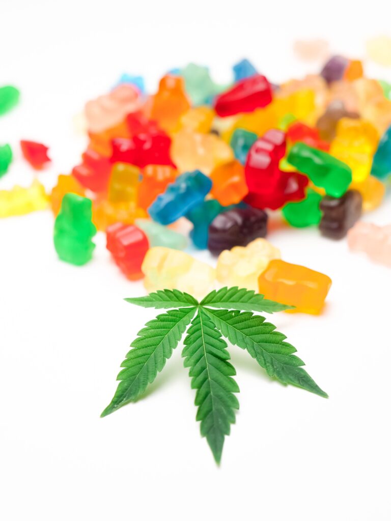 A single green cannabis leaf in the foreground with a blurry pile of colorful gummy bears in the background.
