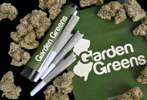 A product layout on a solid black background featuring three white, cone-shaped cannabis pre-rolled joints. The joints are positioned alongside a green storage tube and a green "Garden Greens" package decorated with a white New Jersey state outline. Multiple loose, trichome-covered cannabis buds are scattered around the products.