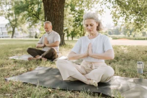 A serene outdoor photograph featuring an older woman and man practicing yoga or meditation in a sunlit park. In the foreground, a woman with grey hair sits cross-legged on a dark yoga mat laid on the grass, her eyes gently closed and hands held at heart center in a prayer position. She wears a loose white t-shirt and cream-colored pants. Behind her to the left, near a large tree, a grey-bearded man sits in the same meditative pose. Soft daylight filters through lush green tree foliage, and a reusable water bottle rests on the grass nearby, creating a peaceful, refreshing atmosphere centered on wellness and mindful aging