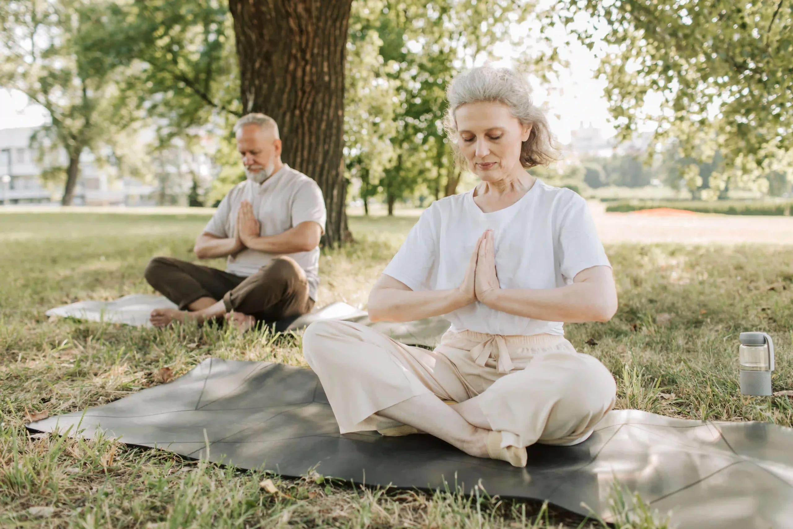 A serene outdoor photograph featuring an older woman and man practicing yoga or meditation in a sunlit park. In the foreground, a woman with grey hair sits cross-legged on a dark yoga mat laid on the grass, her eyes gently closed and hands held at heart center in a prayer position. She wears a loose white t-shirt and cream-colored pants. Behind her to the left, near a large tree, a grey-bearded man sits in the same meditative pose. Soft daylight filters through lush green tree foliage, and a reusable water bottle rests on the grass nearby, creating a peaceful, refreshing atmosphere centered on wellness and mindful aging