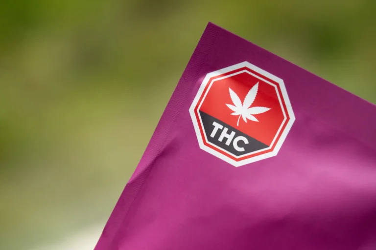 A close-up, diagonal photograph focusing on the top corner of a matte magenta product pouch, set against a softly blurred, vibrant green background. Prominently printed on the packaging is the official Canadian standardized cannabis warning label—a red octagon bordered in white, featuring a stylized white cannabis leaf above a horizontal black banner with the letters 'THC' in bold white text. The clean macro shot highlights regulatory compliance, product safety, and legal cannabis packaging design.