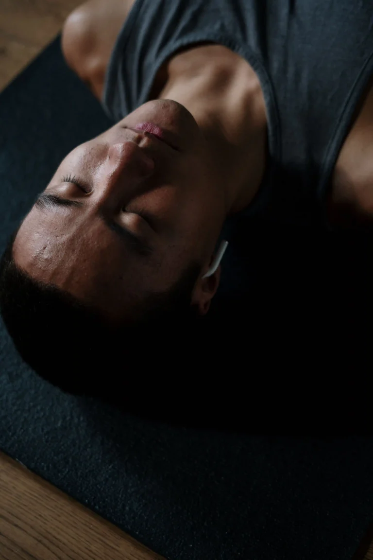 A top-down, close-up photograph of a man lying flat on his back on a dark, textured yoga mat, capturing a moment of deep relaxation, meditation, or rest. His eyes are softly closed with a calm, peaceful expression, and a single white wireless earbud is visible in his ear. He is wearing a simple grey sleeveless tank top. The moody, low-key lighting accentuates his facial features and creates soft shadows, beautifully symbolizing mindfulness, wellness, audio-guided meditation, or a restful savasana yoga pose at home.