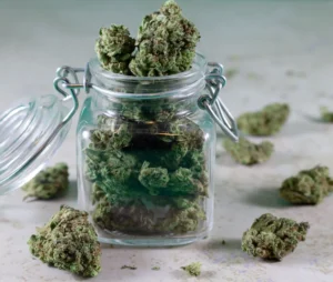 A close-up studio photograph featuring a small, clear glass storage jar with a silver metal flip-top latch filled to overflowing with cured cannabis buds. The open lid leans to the left while several dense, frost-covered green buds spill over the top edge. Additional dried flower buds are scattered across the surrounding light-grey textured surface against a softly blurred background, creating a clean and professional product display