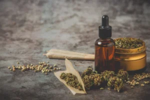 An atmospheric, eye-level photograph displaying a variety of cannabis and hemp products artfully arranged on a textured grey stone surface, ideal for wellness, medicinal, or legal dispensary content. On the right, a small amber glass dropper bottle with a black cap stands beside a open metallic gold herb grinder filled with finely ground flower. In the foreground, a open rolling paper holding ground cannabis sits next to several dried green buds. To the left, two pre-rolled joints lie horizontally in the mid-ground, while small, speckled hemp seeds are scattered loosely across the stone tabletop. The moody, warm lighting emphasizes the natural textures and organic tones of the elements.