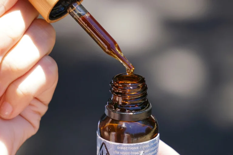 A close-up, sunlit photograph capturing a hand holding a glass dropper filled with dark amber oil suspended just above the neck of an open amber glass tincture bottle. A single golden droplet hangs delicately from the tip of the glass pipette. The bottle features a wooden-capped dropper top and a partially visible label displaying printed text that reads 'DIRECTIONS / PEEL TO OPEN' alongside cannabis product details. Natural sunlight illuminates the rich, viscous liquid and highlights the glass textures against a softly blurred background.