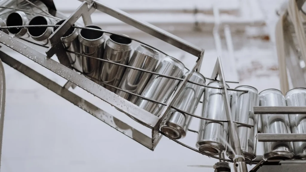A close-up, industrial photograph of multiple silver aluminum cans moving along a metallic conveyor system or guided track within a manufacturing plant. The cans, in various horizontal and vertical stacks, appear raw and unpainted. They are guided by metal rails and a wire frame structure. The cans show some moisture or residue, and the factory environment with pipes and a blurry ceiling is visible in the background.