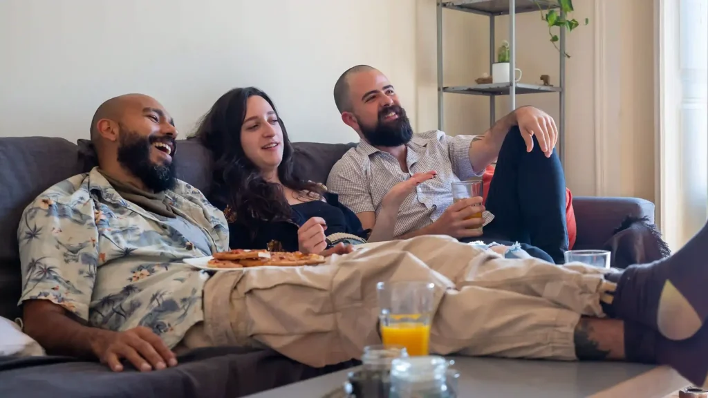 Three friends laughing on a couch while sharing pizza, drinks, and cannabis at a relaxed at-home hangout